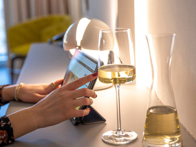 Hände bedienen ein Tablet, mit einem Glas Weiswein daneben stehend.